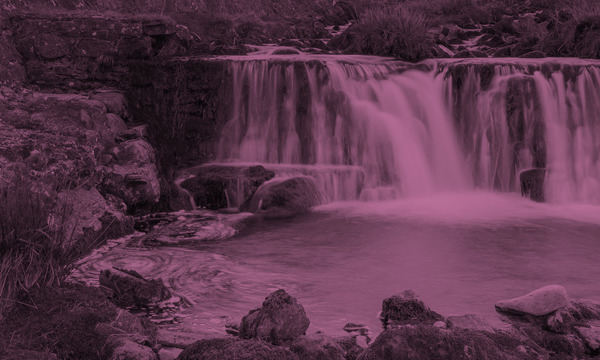 Waterfall cascading over rocks into  pool, surrounded by green hills and mountains.