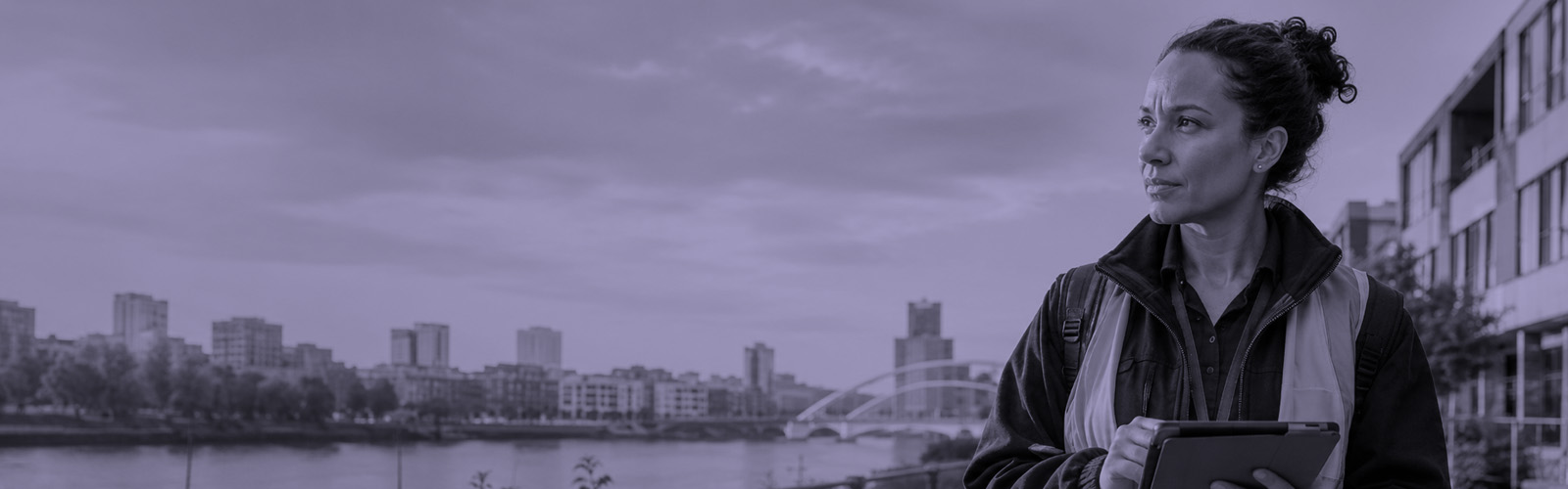 A woman in a reflective vest holds a tablet while standing outdoors near a river, with city buildings and a bridge in the background